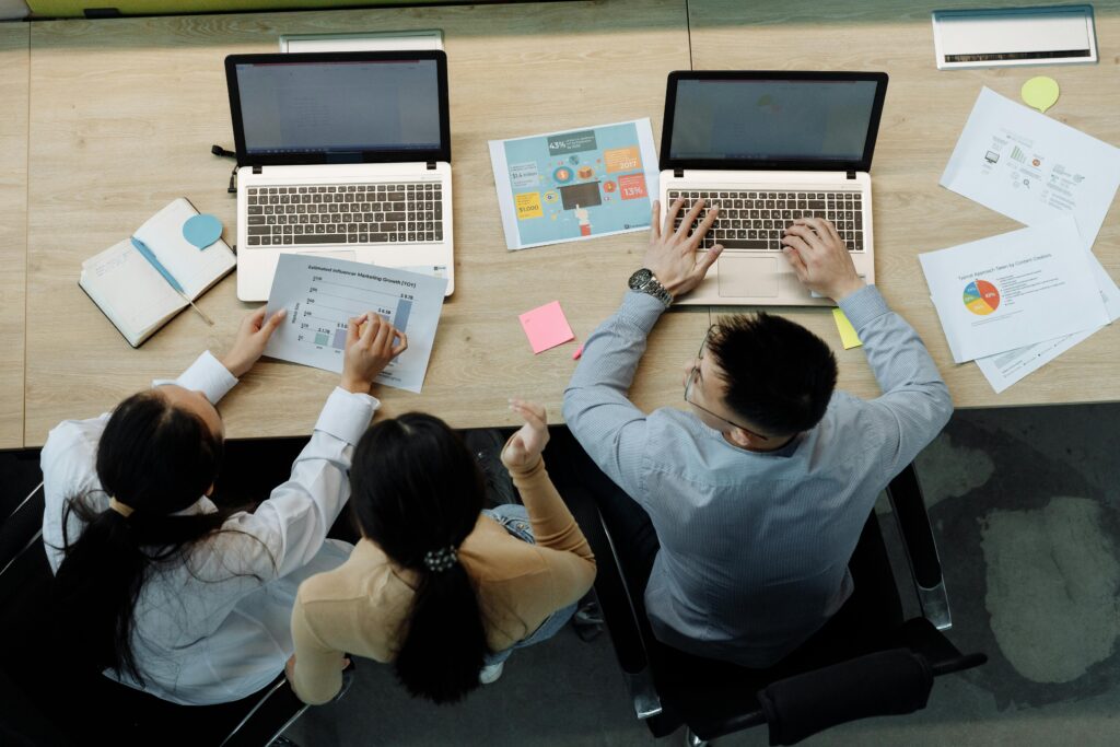Three colleagues working on laptops and documents at a desk, collaborating on business projects.